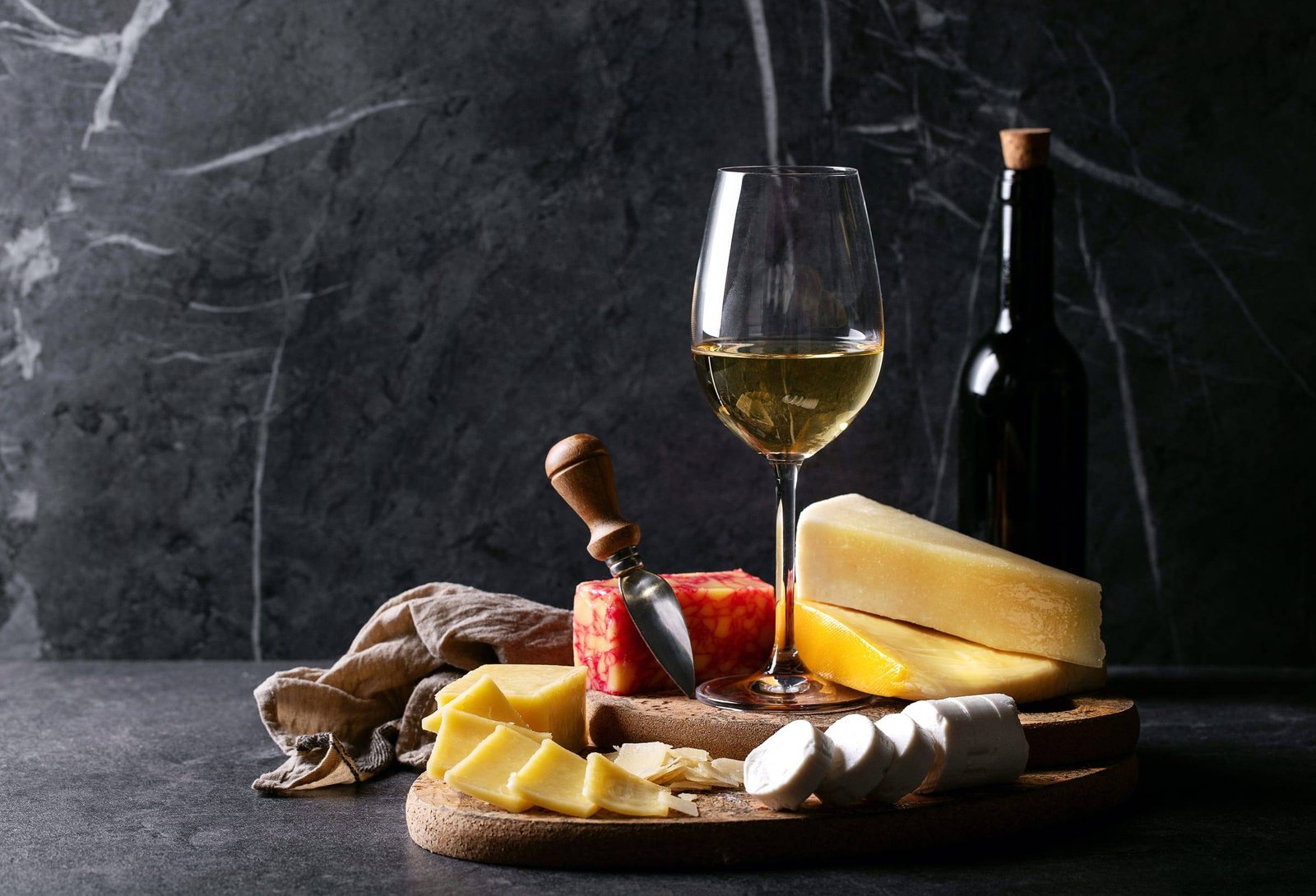 Plateau de fromages, verre de vin blanc et bouteille sur fond sombre